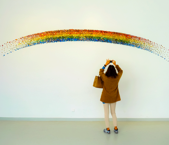 Person in art gallery, facing a glitter rainbow mural.